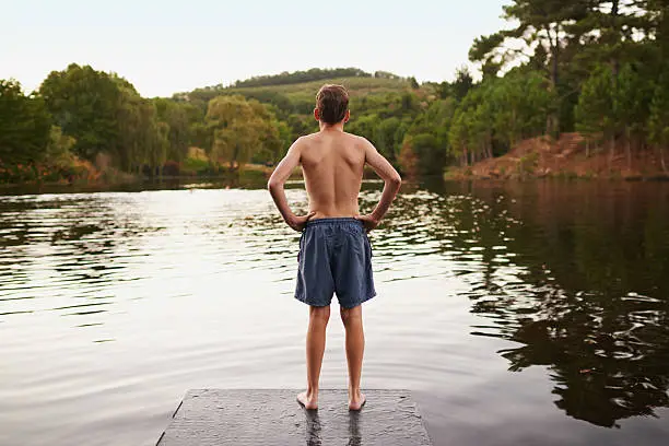 A teenage boy standing on the edge of a pier and looking out over a lake