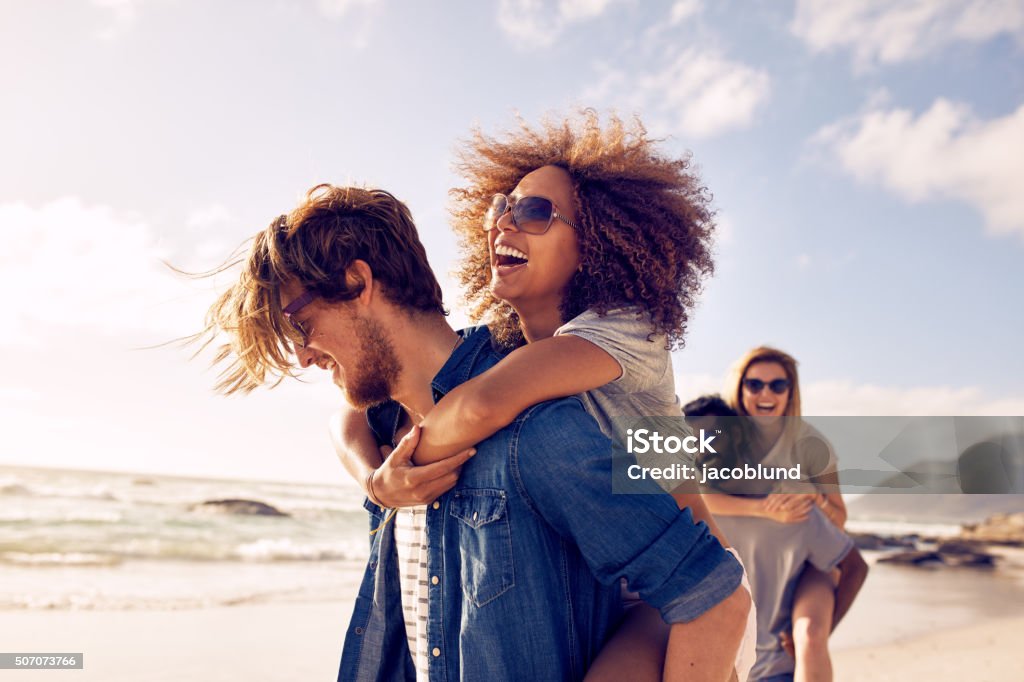 Young friends enjoying a day at beach. Group of friends walking along the beach, with men giving piggyback ride to girlfriends. Happy young friends enjoying a day at beach. Friendship Stock Photo