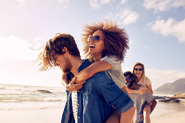 Young friends enjoying a day at beach. Group of friends walking along the beach, with men giving piggyback ride to girlfriends. Happy young friends enjoying a day at beach. summer fun stock pictures, royalty-free photos & images