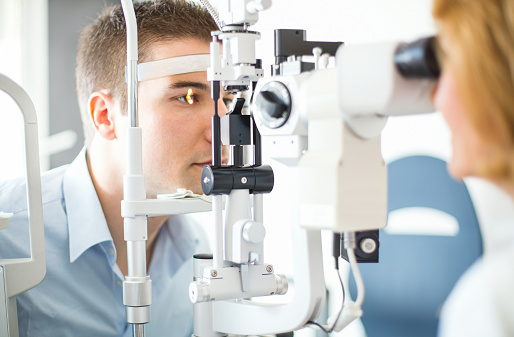 Middle aged caucasian man having her eyes examined at the optician.His head is placed in phoropter apparatus while senior female doctor is examining his retina. The man has short brown hair and light blue shirt.