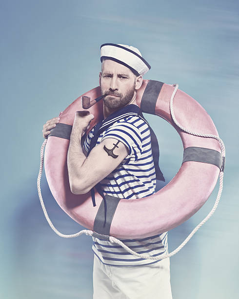 Bearded sailor holding lifebuoy and smoking pipe Summer portrait of bearded sailor man wearing white and blue striped t-shirt and sailor hat, holding lifebuoy on shoulder, flexing her arm with tatoo on shoulder and smoking pipe. Standing against blue background. Studio shot, one person.  shoulder tattoo designs for men stock pictures, royalty-free photos & images
