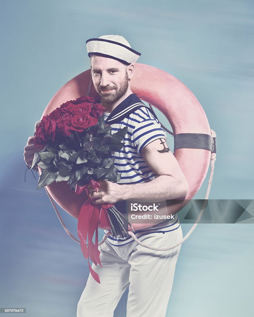 Bearded sailor holding lifebuoy and bunch of roses Summer portrait of bearded sailor man wearing white and blue striped t-shirt and sailor hat, holding lifebuoy on shoulder and bunch of roses in hand, smiling at camera. Standing against blue background. Studio shot, one person.  Sailor Stock Photo