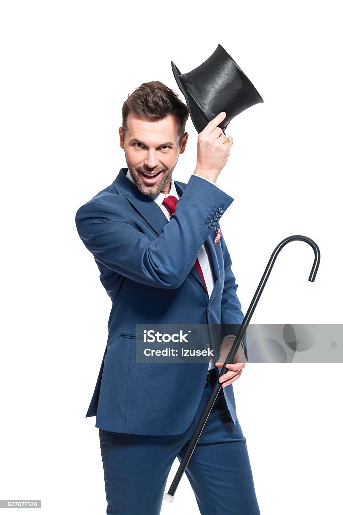 Charming businessman wearing suit and cylinder hat Portrait of elegant businessman wearing suit and cylinder hat, holding walking cane, smiling at the camera. Studio shot, one person, isolated on white. Magician Stock Photo
