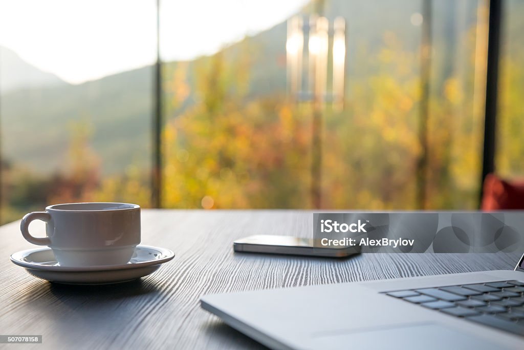 Business Travel Morning Composition with Computer Coffee and Telephone Business Travel Composition with cropped Computer Coffee Mug and Telephone on black grey Wood Table and large Windows Garden and Mountains on Background Nature Stock Photo