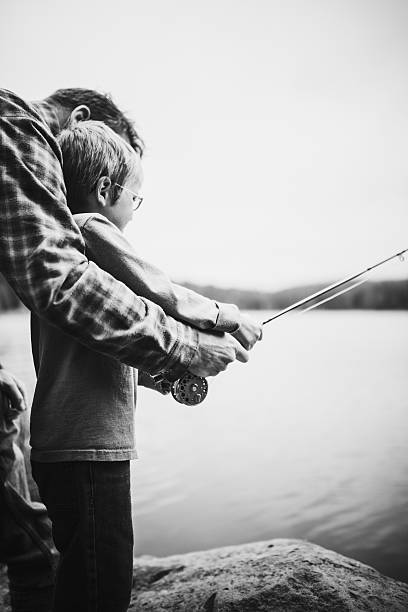 A grandfather is teaching his grandson to fish during summer. They are both standing on the dock. It is a beautiful summer day at sunset. Across the lake, there is a mountain, Quebec, Canada.