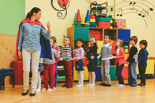 Group of children playing with their teacher in kindergarten. Kids are standing in a row and smiling. Teacher is on the front of a row holding thumb up. Teacher wearing super hero cape.