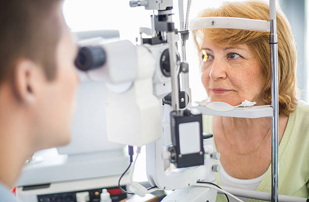 Senior woman visiting optician. Senior caucasian woman having her eyes examined at the optician.Her head is placed in phoropter apparatus while middle aged male doctor is examining her retina. The woman has mid length yellow brown hair and wearing light breen blouse. eye exam stock pictures, royalty-free photos & images