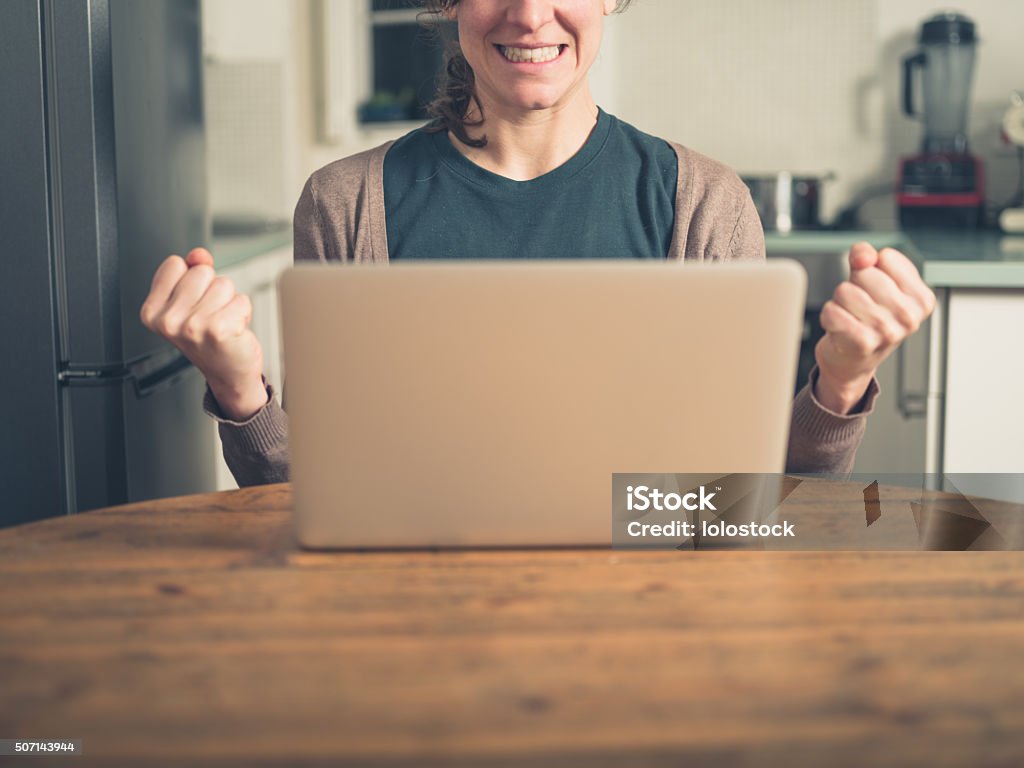 Young woman with laptop fist pumping in kitchen A young woman is using her laptop in a domestic kitchen and is doing a double  fist pump as she is getting excited Achievement Stock Photo