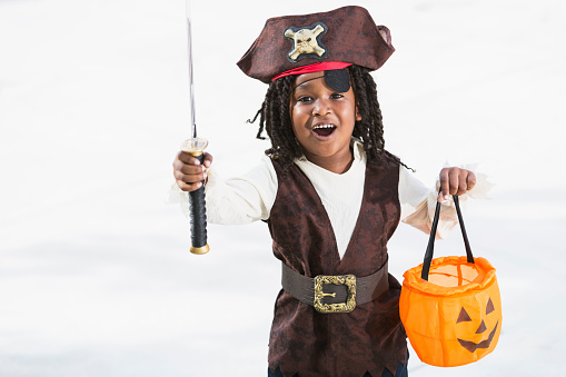 African American boy (6 years) wearing pirate halloween costume, holding jack o lantern treat bag, excited about going trick or treating.