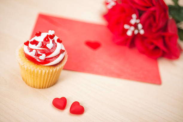 Valentine's Day strawberry shortcake cupcake with heart shaped sprinkles, a card in a red envelope with two red heart shape candies and red roses in the background.