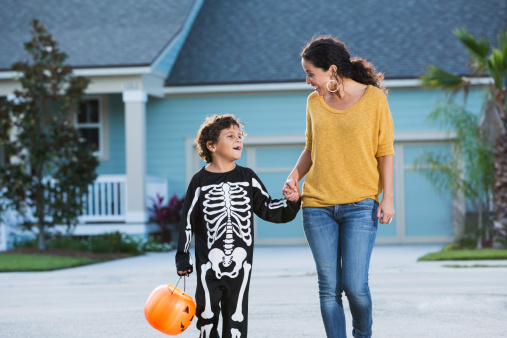 Hispanic woman (40s) with son (6 years) on halloween wearing skeleton costume, walking on street in front of house, smiling, holding hands and talking.