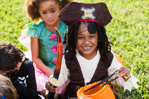 African American boy (6 years) dressed in pirate costume for halloween, sitting with group of children outdoors on grass.