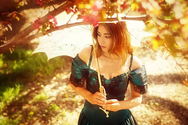 Horizontal colour garden shot of young woman in green ball gown. Overhead view looking down through branches of flowering crab apple. She's hodling a white lace parasol and looking to her right, viewer's left.