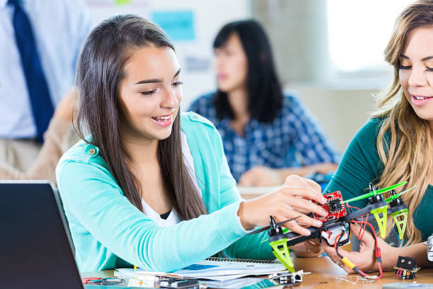 Female high school students build drone at STEM school Two female STEM high school students build drone together in class or after school engineering club. One student holds the drone while the other assembles a part. They are having fun and smiling as they work together. A laptop is on the table. The students are dressed casually. Teachers and students are in the background working on projects. high school high school student science multi ethnic group stock pictures, royalty-free photos & images