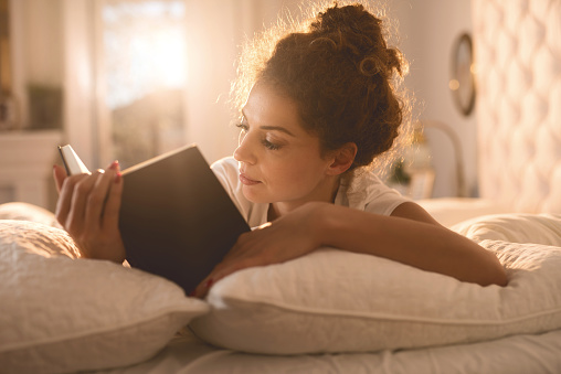 Young woman lying in bed and reading a book.