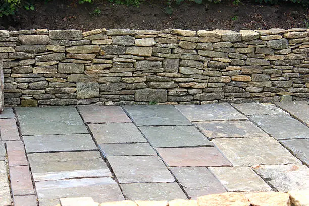 Photo showing a large sunken garden under construction, with the dry stone walls / raised beds being pictured partially built during this hardscaping phase.  The flower beds will be landscaped with Alpine plants, rocks and scree gravel as soon as the walls are finished.  A large area of flagstone paving slabs has been laid to make the garden accessible for disabled people using wheelchairs.