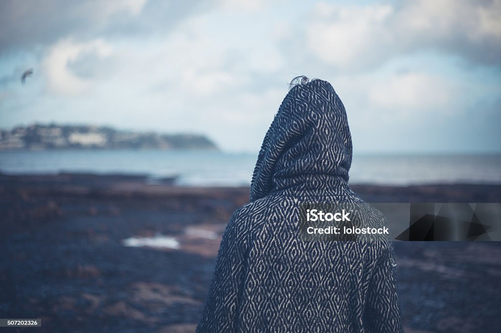 Young woman in hooded coat walking on beach A young woman wearing a hooded coat is walking on a dramatic beach in the winter Sadness Stock Photo