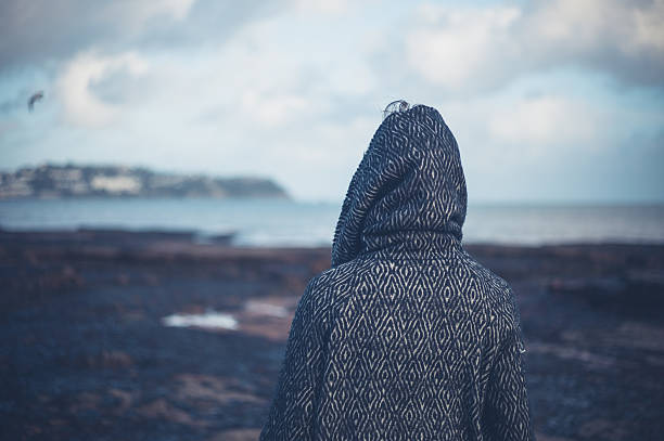 Young woman in hooded coat walking on beach A young woman wearing a hooded coat is walking on a dramatic beach in the winter walking beach women sadness stock pictures, royalty-free photos & images