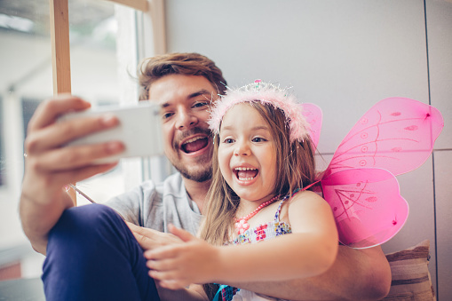 Father and daughter enjoying at home. Little girl wearing fairy costume and tiara. Sitting on window wooden seat. Taking selfies with smart phone.
