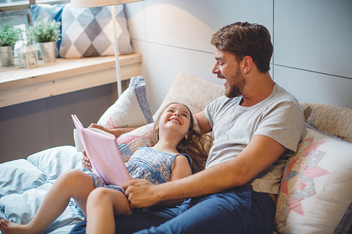 Father and daughter enjoying at home. Sitting on bed and reading book together.