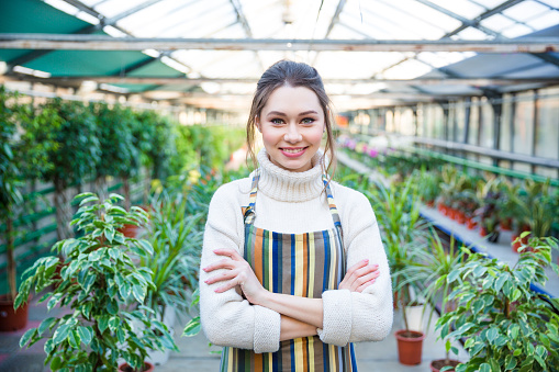 Beautiful happy young woman gardener in white sweater and colorful striped apron standing in orangery