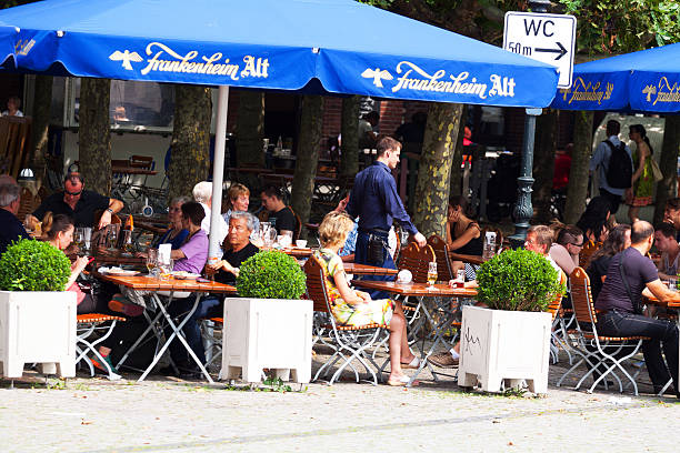 Düsseldorf, Germany - July 22, 2014: People are sitting at tables of bistro and beer garden on square in Altstadt of Düsseldorf in summer. There are young, adult and senior people. Waiter is walking between tables. He is holding a tray in his hand. Scene is at square Burgplatz.