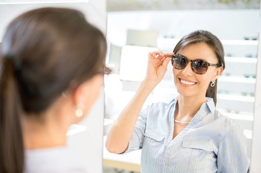 Happy woman buying sunglasses and looking at the mirror at the optician's shop for the summer - lifestyle concepts