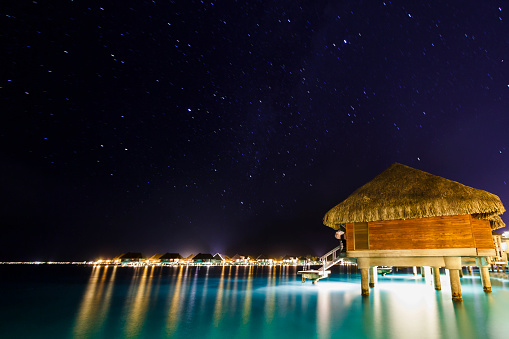 Bora Bora, French Polynesia - August 15, 2014: A DSLR long exposure photo taken from the deck of a over water bungalow in Intercontinental Bora Bora Resort  and Thalasso Spa. It is a clear night and there are many stars shining in the sky. The flat water suface of the lagoon is reflecting the colorful lights from the many bungalows of the resort.