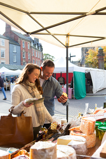 Man smiles as he picks up some cheese that on sale on a food market stall. He stands close to a woman as they both look at what he is holding in his hand. The sheet of the outdoor market stall can be seen above their heads and a large selection of various cheese on show in front of them.