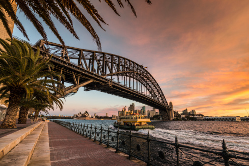 Sydney Harbour Bridge with Ferry, Opera House and Palmtrees at Dusk. Twilight warm glow in the sky.