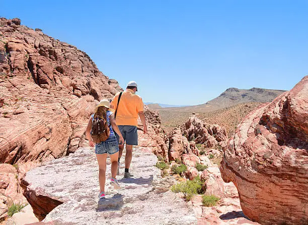Father and daughter hiking in Red Rock Canyon, Nevada, USA. Girl is wearing a backpack. Blue sky in the background.