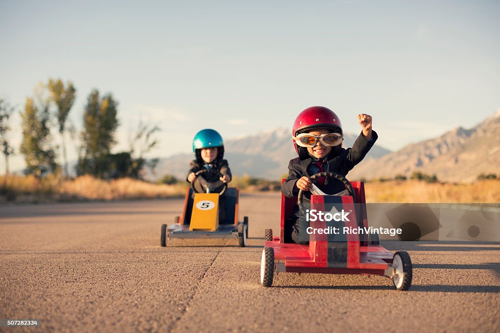 Young Business Boys in Suits Race Toy Cars A young boy dressed as a businessman raises his arm in success as his homemade box car is in first place. Both boys are wearing helmets and goggles.  Child Stock Photo
