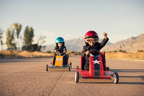 Young Business Boys in Suits Race Toy Cars A young boy dressed as a businessman raises his arm in success as his homemade box car is in first place. Both boys are wearing helmets and goggles.  car image stock pictures, royalty-free photos & images