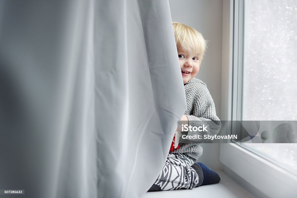 Toddler boy playing at home Toddler boy playing on the window sill at home Hide And Seek Stock Photo