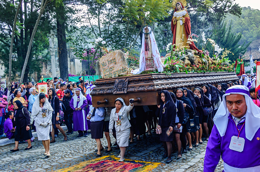 Antigua, Guatemala - April 3, 2015: Devout Catholic locals participate in Good Friday procession. Women carry float with Virgin Mary over handmade dyed sawdust carpet (alfombra) in Spanish colonial town & UNESCO World Heritage Site with most famous Holy Week celebrations in Latin America. Purple robed men (cucuruchos) carry float with figure of Jesus. Locals & tourists watch.