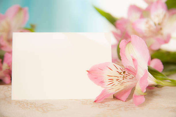 Easter or Mother's Day.  A bouquet of pink Alstroemeria flowers lies behind and to the side of a blank white notecard for your copy.  No people. Blue window background. Feminine scene.