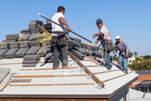 A roofer holds nails in his mouth as he is handed an eave riser that he is getting ready to put on a new roof.