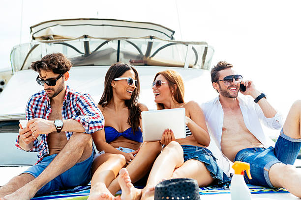 Cheerful Friends Using Their Gadgets On Yacht. Cheerful Friends Yachting Together and having fun. Sunbathing and talking on a deck. Girls Making selfie and using digital tablet. One man texting on his smart phone and other man talking on the phone. yacht selfie stock pictures, royalty-free photos & images