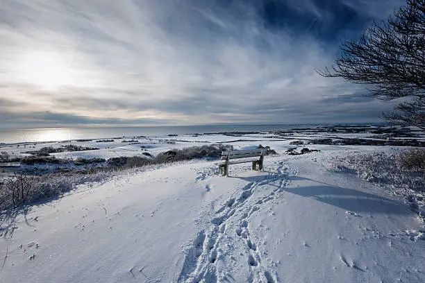 Winter view over the snow covered island of Møn in Denmark. Taken on Kongsbjerg, the highest part of the island and  looking over towards the town of Stege and the Baltic beyond.  Colour, horizontal format, with lots of copy space. The wooden bench in the foreground is known as a "tænkebæk" or "thinking bench". They have been  put up on some of the most picturesque spots all over the island of Møn in Denmark.