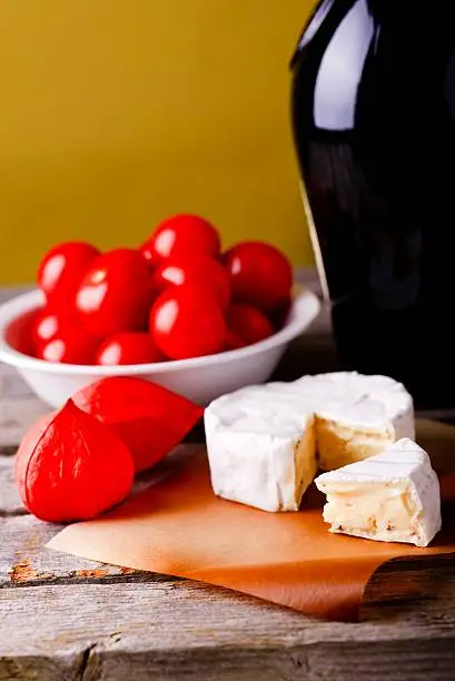 Vertical photo of camembert cheese with herbs placed on piece of paper and old worn wooden table with few physalis blooms, white bowl with tomatoes and glass of red wine.