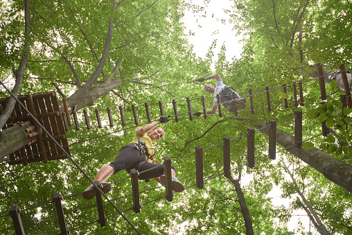low angle view of two people in adventure park climbing the wooden bridge and enjoying sport activities.