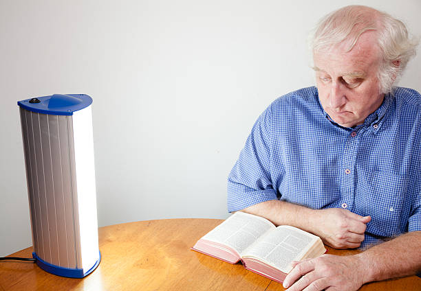 senior man reads by seasonal affective disorder SAD lamp senior man reads by seasonal affective disorder SAD lamp - these lamps simulate strong daylight and are believed to be effective i controlling  depression associated with lack of daylight in winter months -  "Seasonal Affective Disorder" or SAD light therapy stock pictures, royalty-free photos & images