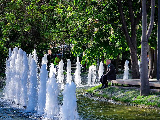 Ankara, Turkey - April 30, 2013:  Old men sitting on a bench is looking a fountain with water games. The men look very absorbed in his own thinking and is trying to get positive thinking from the nature he is looking at. The man is well dressed an looks like that he is still working and he just take few minutes to relax. The light is coming from an overcast day anf filtering  through the trees leaves.