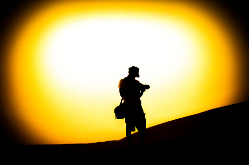Silhouette of a young photographer walking on the dunes of Dubai. A creative composition with high contrast background showcasing bright sunset, vignetted image.