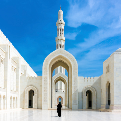 Woman in black Abaya Gown walking towards the giant arch in the famous Sultan Qaboos Grand Mosque in Muscat, Oman, Middle East, Arabia.