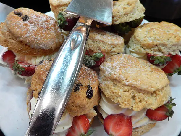 Photo showing an indulgent plate of luxury homemade cream scones, ready for a cream tea at an afternoon tea party.  The scones are pictured with a silver cake slice and displayed on a white plate.  A traditional cream tea (also referred to as a Devonshire, Devon or Cornish cream tea) consists of scones and clotted cream.
