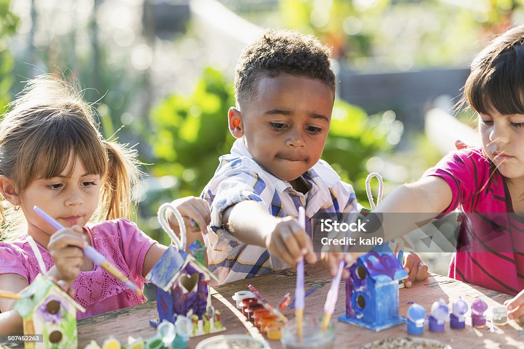 Multi-ethnic children painting bird houses outdoors A multiracial group of three serious young children doing an arts and crafts project, painting little wooden bird houses for Earth Day.  It is a bright, sunny day and they are sitting at a wooden table reaching with their paintbrushes.  An African American boy, 5 years old, is sitting in the middle between two Hispanic girls. Child Stock Photo