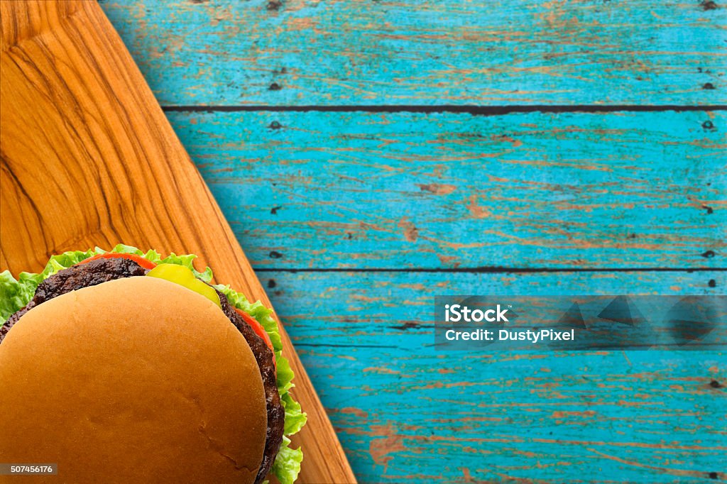 Dressed Burger on Blue Picnic Table Dressed burger with lettuce, tomatoes, and pickles rests on cutting board with a rustic blue picnic table background Beef Stock Photo