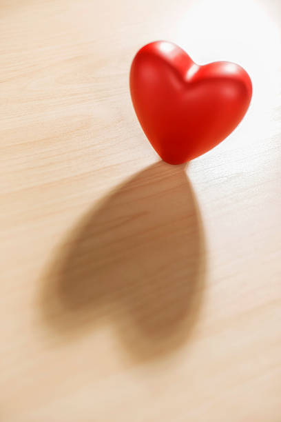 A single red heart casting a heart shaped shadow on a light colored wood table for Valentine's day