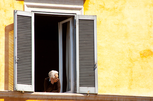 Rome, Italy - May 1, 2009: A senior woman leans out a tall window of a building with a sunlit mottled yellow wall.